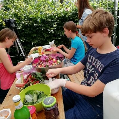 Bild vergrößern: Kinder sitzen an einer Bierzeltgarnitur und verarbeiten Obst- und Gemüse zu natürlichen Farbstoffen